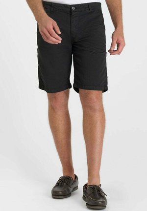 Man wearing black knee-length shorts and dark brown leather lace-up loafers, standing against a plain white background.