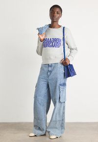 Grå sweatshirt med lilla bokstaver, brede blå denim cargo bukser og en liten blå håndveske. Modellen holder et denimstykke over skulderen.