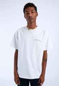 Bílý bavlněný tričko s malým zeleným grafickým textem "DUMP GOOD TIMES" na hrudi. Model nosí vrstvené korálkové náhrdelníky.