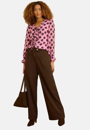 Woman wearing pink blouse with large brown polka dots, brown wide-leg trousers, brown heeled shoes, holding a brown handbag.