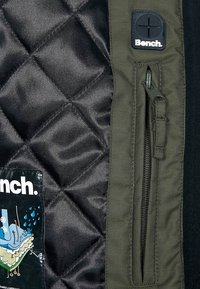 Tessuto nero trapuntato con dettagli verde oliva, dotato di una tasca con zip e un'etichetta con il logo Bench, che mette in mostra una texture liscia e un design strutturato.