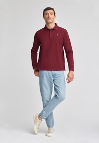 Burgunderfarbendes Langarm-Poloshirt mit Kragen, kombiniert mit hellblauen Jeans und beigen Sneakers; verfügt über ein kleines Logo auf der Brust.