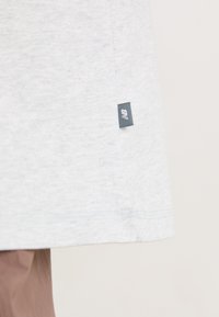 T-shirt en coton gris clair avec une étiquette latérale portant un logo noir. Texture douce, col rond standard et manches courtes.