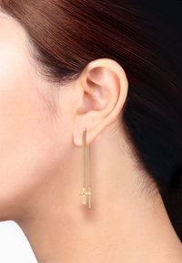 Goldene Ohrringe mit Ketten, die zwei hängende Kreuzformen zeigen, mit einem polierten Finish und feinen Details, getragen am Ohr vor einem neutralen Hintergrund.