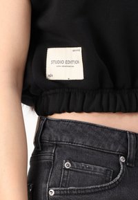 Juodas trumpas topas su elastingu apatine dalimi, puošiamas baltu etiketu su užrašu "STUDIO EDITION", ir pilkos aukšto liemens džinsai su sidabrinėmis sagomis bei siuvinėjimu.