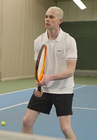 Λευκό πόλο μπλουζάκι με μαύρες λεπτομέρειες, μαύρα σορτς και κρατώντας μια πορτοκαλί και μαύρη ρακέτα τένις, τοποθετημένος σε μπλε εσωτερικό γήπεδο.