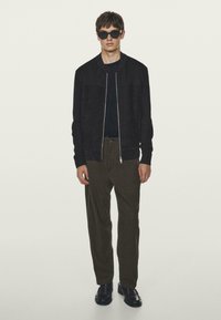 Giacca bomber nera con chiusura a zip, abbinata a pantaloni verde oliva scuro. Il modello indossa occhiali da sole neri e scarpe nere. Texture liscia in tutto.