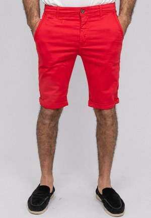 Homme portant un short rouge vif retroussé, les mains dans les poches et des chaussures noires à enfiler, sur un fond uni.
