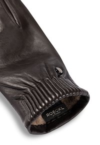 Nahaufnahme eines schwarzen Lederhandschuhs mit elastischem Bündchen, einem kleinen Knopf und einem Stoffetikett mit der Aufschrift "ROECKL MÜNCHEN."