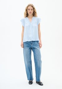 Blusa azzurro chiaro a maniche corte con colletto, abbinata a jeans blu larghi e scarpe nere, su uno sfondo bianco.