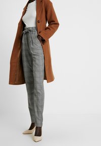 Bruine wollen jas met zwarte knopen, witte geribbelde top, grijze geruite hoog getailleerde broek en beige puntige hakken.