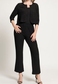 Czarny kombinezon z błyszczącą teksturą, z krótką kurtką, rękawami o długości trzy czwarte oraz spodniami o luźnym kroju. W połączeniu z czarnymi szpilkami.