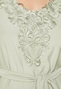 Vestido verde claro com bordado intrincado em padrão floral à volta do decote, acompanhado de um cinto amarrado na cintura para um efeito ajustado.