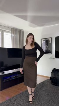 Juodas ilgarankovinis viršus su kvadratine iškirpte, derinamas su ruda iki kelių siekiančia sijonu ir juodais aukštakulniais su dirželiais. Neutralus patalpos fonas.