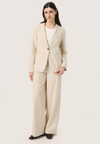 Beige kostym med en figursydd kavaj med så kallade notch-lapel, en svart knapp fram, och vida byxor; slätt tyg med minimala texturdetaljer.