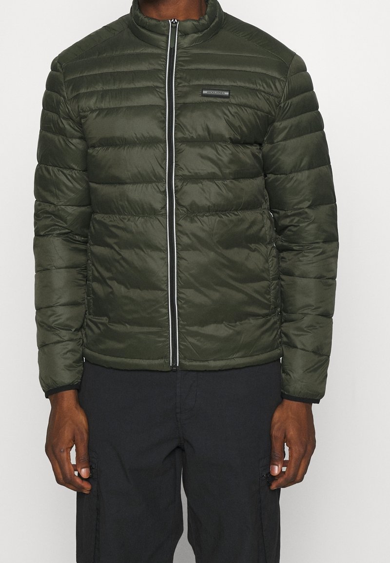 Olivgrön puffjacka med horisontell sömnad, hög krage, framdragkedja och logopatch på vänster bröst. Slät, lättviktig material.