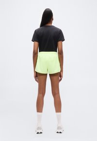 Μαύρο μπλουζάκι συνδυασμένο με νεον πράσινα σορτς με ελαστική μέση και αθλητικά παπούτσια, πάνω σε απλό λευκό φόντο.