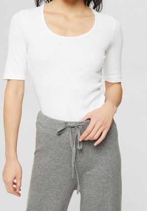 Femme portant un haut blanc ajusté à manches courtes et un pantalon en maille gris à taille coulissante, debout devant un fond uni.