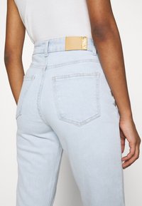 Jeans de tiro alto en azul claro con bolsillos traseros, que presentan una etiqueta de cuero marrón en la cintura con la inscripción "ONLY" en letras doradas.