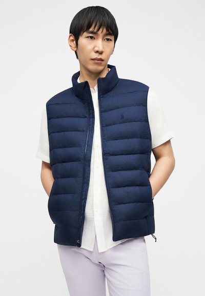 Άνδρας που φοράει μπλε σκούρο αμάνικο μπουφάν με ραφή πάνω από λευκό πουκάμισο με κοντά μανίκια και ανοιχτό μωβ παντελόνι, με τα χέρια στις τσέπες του μπουφάν.