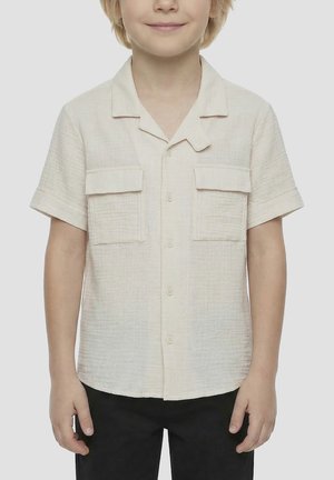 Niño con una camisa de manga corta color beige claro con dos bolsillos en el pecho y pantalones negros, de pie frente a un fondo liso.