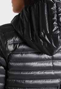 Chaqueta de plumas negra con capucha, diseño acolchado horizontal, textura brillante y acentos elásticos en el cuello y la capucha.