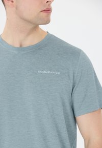 Mies, joka on pukeutunut vaaleansiniseen urheilupaitaan, jossa on "ENDURANCE"-logo, katsoo sivulle valkoista taustaa vasten.