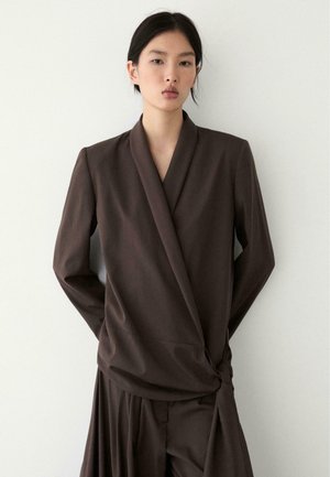 Frau trägt ein dunkelbraunes Wickelblazer mit seitlicher Schleife und passendem plissee Rock, steht vor einem schlichten weißen Hintergrund.