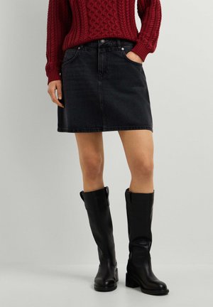 Person trägt einen dunklen Jeansrock, roten Strickpullover und kniehohe schwarze Lederstiefel und steht vor einem schlichten Hintergrund.
