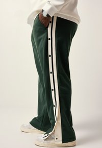 Green sweatpants with side snap-button closure, contrasting white and black stripes, and a smooth fabric texture. Worn with light sneakers.