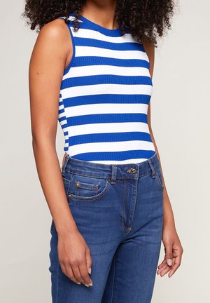 Woman wearing blue and white horizontal striped sleeveless top and blue high-waisted jeans, standing against a plain background.