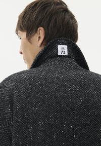 Uomo con capelli corti castani che indossa un cappotto a spina di pesce di colore grigio scuro con il colletto rialzato, che mostra un'etichetta bianca con scritto "M 73."