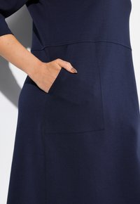 Navyblaues Kleid aus weichem Stoff mit rundem Ausschnitt, dreiviertellangen Ärmeln und einer Seiten Tasche mit kontrastierenden Nähdetails.