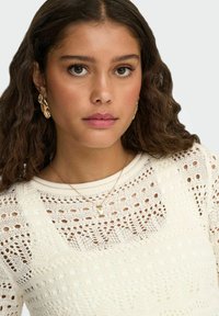 Pull en tricot avec un motif texturé en crème, doté d'un col large. Boucles d'oreilles en or et un collier délicat en chaîne d'or ornent le modèle.