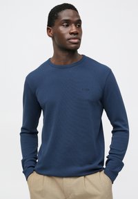 Camisola azul-marinho de manga comprida feita de tecido texturizado, com decote redondo e logótipo discreto da marca no peito, combinada com calças bege.