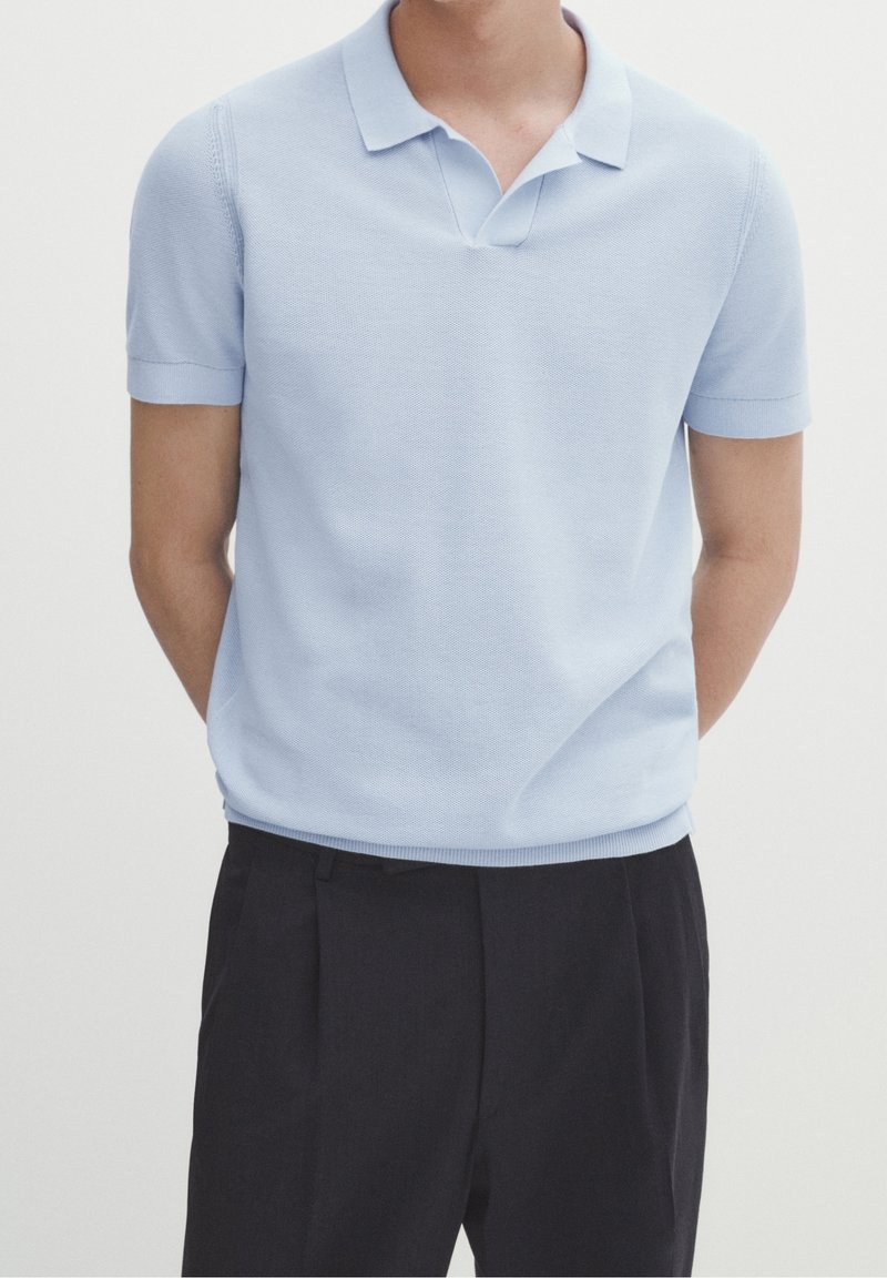 Homme portant un polo en maille bleu clair à manches courtes, rentré dans un pantalon noir plissé, les mains derrière le dos.