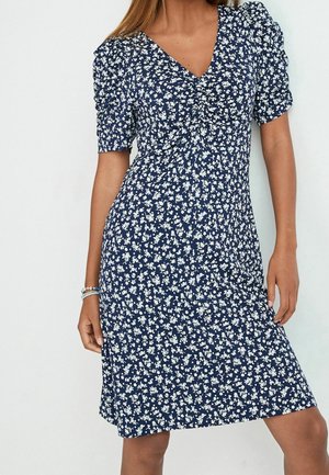 Femme portant une robe bleu marine à imprimé floral blanc, manches courtes bouffantes et encolure en V, debout devant un fond uni.