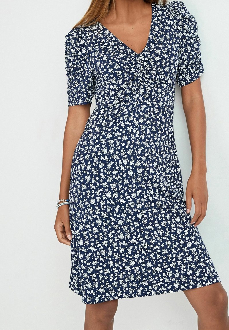 Femme portant une robe bleu marine à imprimé floral blanc, manches courtes bouffantes et encolure en V, debout devant un fond uni.