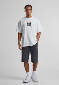 Weißes übergroßes T-Shirt mit schwarzem Grafikdruck, kombiniert mit anthrazitfarbenen Denim-Shorts und weißen Sneakern. Lässiger Schnitt und strukturierter Stoff.