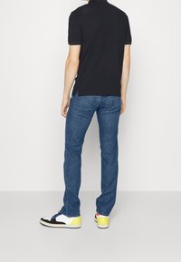 Černé polo tričko s krátkým rukávem spojené s modrými džínami z denimu. Tenisky mají bílé, černé a žluté detaily. Pohled zezadu.