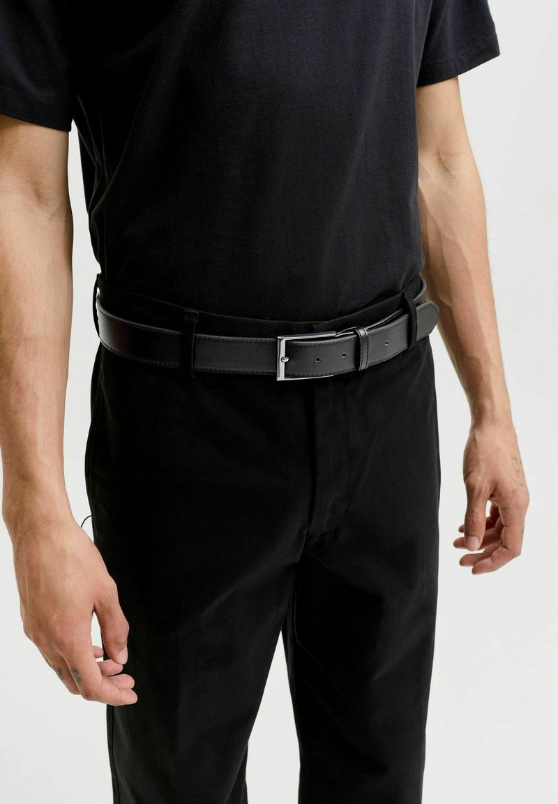 Homme portant un pantalon noir, une ceinture noire avec une boucle argentée et un t-shirt noir, debout les bras détendus le long du corps devant un fond uni.