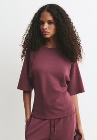 Burgundské sportovní tričko s krátkými rukávy, které má volný střih a hladkou texturu. Švy podél boků zvýrazňují design.