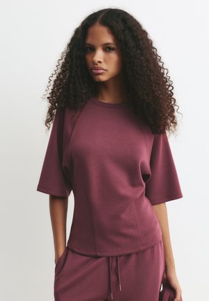 Haut de sport bordeaux à manches courtes, avec une coupe décontractée et une texture lisse. Les coutures sur les côtés rehaussent le design.