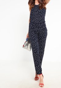 Námořnický overal s bílými puntíky, bez rukávů, s elastickým pasem a zúženými nohavicemi. V kombinaci s červenými sandály a metalickou kabelkou.