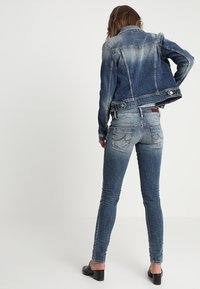 Jeansowa kurtka w ciemnym niebieskim kolorze, przechodząca w jaśniejsze odcienie. Dopasowane jeansy z tylnymi kieszeniami i detalami przeszyć. Widoczne czarne buty na obcasie.