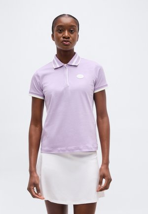 Femeie purtând un tricou polo violet deschis cu margine albă și o fustă albă, stând în picioare pe un fundal simplu, deschis.