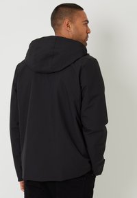 Sort vandtæt jakke med hætte, der har en glat overflade, minimalistisk design og en let forlænget bagkant.