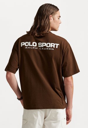 Bărbat cu păr brown până la umeri, purtând un tricou Polo Sport Ralph Lauren maro și pantaloni albi, văzut din spate.