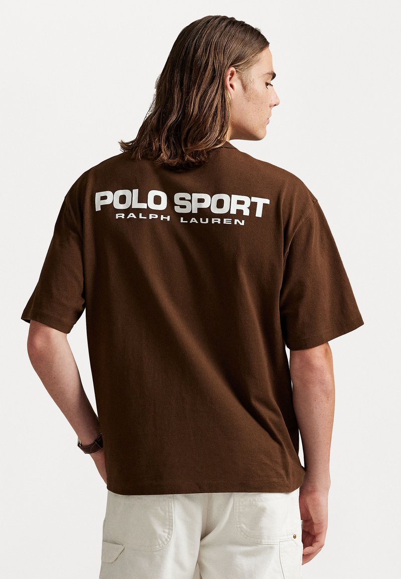 Vīrietis ar plecu garumu brūniem matiem, ģērbts brūnā Polo Sport Ralph Lauren T-kreklā un baltās biksēs, skatīts no aizmugures.