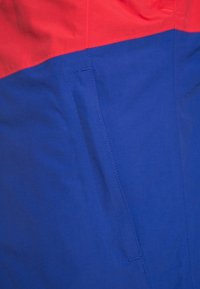 Tessuto a blocchi di colore con una sezione superiore rossa e una sezione inferiore blu. Presenta una tasca nascosta con dettagli di cucitura semplici. Texture liscia.